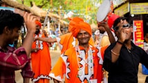 A group of people in vibrant traditional attire, including bright orange turbans and robes, are participating in a lively celebration. One individual is holding a tambourine, while others around him appear to be dancing and enjoying festive music. The setting is an outdoor cultural fair with colorful banners and stalls in the background.