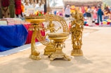 The image features intricately designed brass sculptures placed on the ground in a marketplace. In the foreground, there is a sculpture of a deity with elaborate detailing and a decorative circular backdrop. Textiles and other decorative items are visible in the background, displayed on tables covered with colorful cloths. Several people are blurred in the background, indicating a busy market atmosphere.