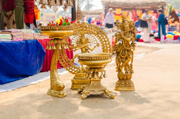 The image features intricately designed brass sculptures placed on the ground in a marketplace. In the foreground, there is a sculpture of a deity with elaborate detailing and a decorative circular backdrop. Textiles and other decorative items are visible in the background, displayed on tables covered with colorful cloths. Several people are blurred in the background, indicating a busy market atmosphere.