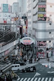 A bustling urban street scene in Japan featuring pedestrians walking along the streets and sidewalks beneath tall commercial buildings with colorful signs and advertisements. A prominent archway sign marks the entrance to a shopping street. Vehicles, including cars and taxis, navigate through a busy intersection below.