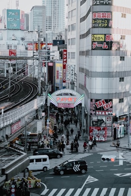 A bustling urban street scene in Japan featuring pedestrians walking along the streets and sidewalks beneath tall commercial buildings with colorful signs and advertisements. A prominent archway sign marks the entrance to a shopping street. Vehicles, including cars and taxis, navigate through a busy intersection below.