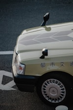 A close-up view of the front-left corner of a car with Japanese text on the side. The car is parked on a textured street surface with visible markings. The car features a light-colored body with detailed geometric patterns on the hood and silver hubcaps.