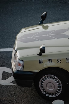 A close-up view of the front-left corner of a car with Japanese text on the side. The car is parked on a textured street surface with visible markings. The car features a light-colored body with detailed geometric patterns on the hood and silver hubcaps.