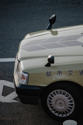A close-up view of the front-left corner of a car with Japanese text on the side. The car is parked on a textured street surface with visible markings. The car features a light-colored body with detailed geometric patterns on the hood and silver hubcaps.