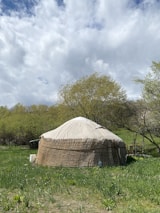 A traditional yurt stands in a lush green meadow surrounded by trees with light green foliage. The sky above is cloudy, with patches of blue visible in between. The landscape suggests a peaceful and serene outdoor setting, possibly in a rural or pastoral area.
