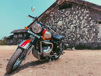 A motorcycle is parked on a dirt ground in front of a stone building. The building has a rustic appearance with a tiled roof and stone masonry walls. The motorcycle is a classic model with chrome details and vibrant colors, prominently featuring a red fuel tank.