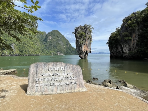 A scenic view of a small island with a tall, narrow limestone rock formation rising from the sea, surrounded by lush green vegetation and clear skies. In the foreground, there is a large stone sign with inscriptions in English and Thai, indicating the site as James Bond Island in Ao Phang Nga National Park. The water is calm, reflecting the natural beauty of the surroundings.