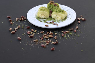A white plate containing a dessert covered with ground pistachios sits on a dark surface. Surrounding the plate, there are scattered whole pistachios and some ground pistachio dust. The dessert appears creamy and is artfully garnished.