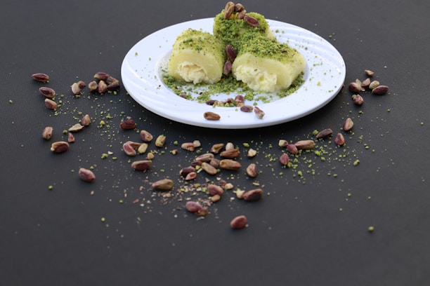 A white plate containing a dessert covered with ground pistachios sits on a dark surface. Surrounding the plate, there are scattered whole pistachios and some ground pistachio dust. The dessert appears creamy and is artfully garnished.