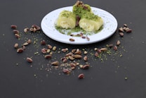 A white plate containing a dessert covered with ground pistachios sits on a dark surface. Surrounding the plate, there are scattered whole pistachios and some ground pistachio dust. The dessert appears creamy and is artfully garnished.