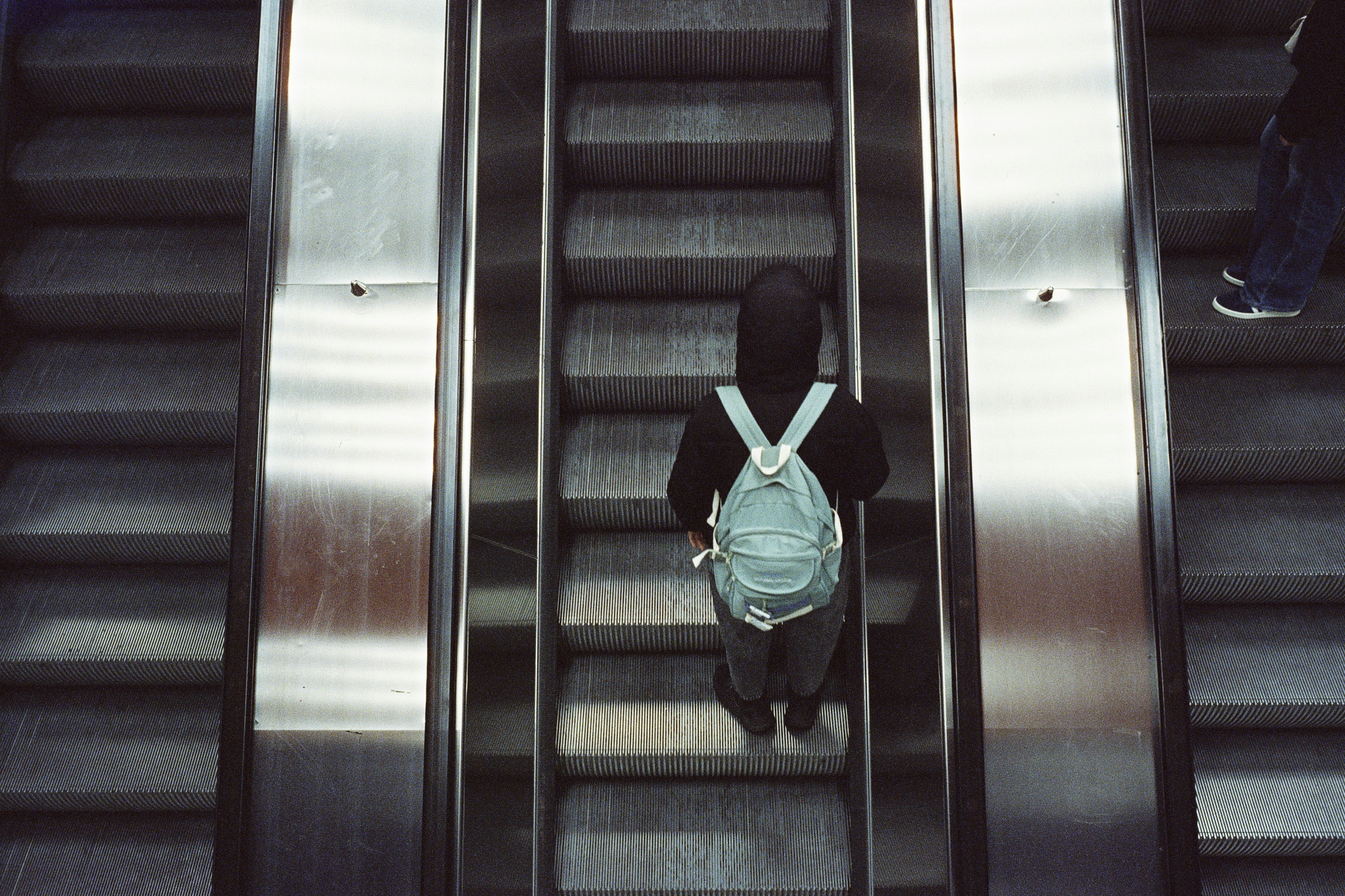 a person on an escalator with a backpack, Local traffic subway station escalator. Leica M6 (1987), Summilux-M 1.4 35mm (1983). Hi-Res analog scan by www.nimmfilm.de – CineStill 800 Tungsten (pushed 1 stopp)