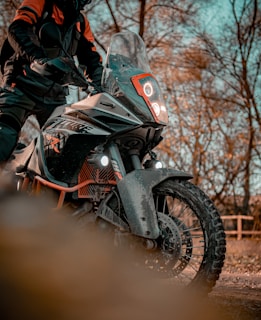 A person in protective gear is standing with a rugged off-road motorcycle. The bike has a distinct orange and black design with prominent headlights and a windshield. The background features tall trees with bare branches, suggesting a fall or early spring setting.