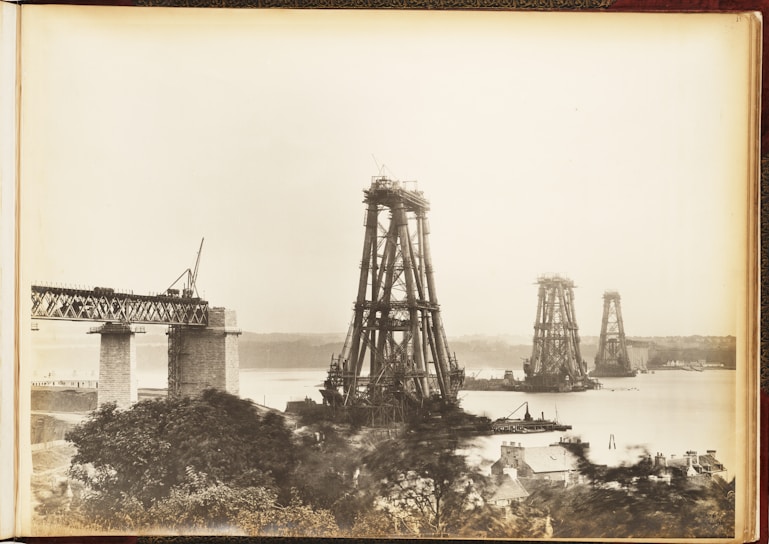 Large metal structures rise from the water, with one partially constructed bridge extending from the left. Scaffolding surrounds the towers, indicating ongoing construction over a body of water. Houses and trees are visible in the foreground.