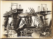 A black and white photograph depicting the construction of a large bridge with significant steel framework. Workers can be seen standing on the bridge structure. The image shows complex architectural scaffolding and hefty metal components, with various construction tools and small buildings nearby.
