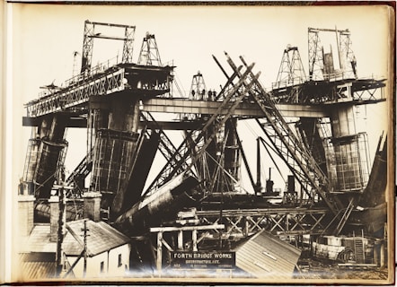 A black and white photograph depicting the construction of a large bridge with significant steel framework. Workers can be seen standing on the bridge structure. The image shows complex architectural scaffolding and hefty metal components, with various construction tools and small buildings nearby.