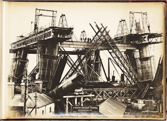 A black and white photograph depicting the construction of a large bridge with significant steel framework. Workers can be seen standing on the bridge structure. The image shows complex architectural scaffolding and hefty metal components, with various construction tools and small buildings nearby.
