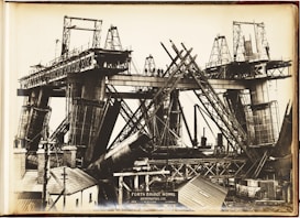 A black and white photograph depicting the construction of a large bridge with significant steel framework. Workers can be seen standing on the bridge structure. The image shows complex architectural scaffolding and hefty metal components, with various construction tools and small buildings nearby.