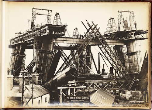 A black and white photograph depicting the construction of a large bridge with significant steel framework. Workers can be seen standing on the bridge structure. The image shows complex architectural scaffolding and hefty metal components, with various construction tools and small buildings nearby.