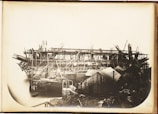 A large industrial structure under construction, featuring a complex framework of scaffolding and beams. Several cranes and wooden structures surround the area, and a body of water is visible beneath the unfinished framework.