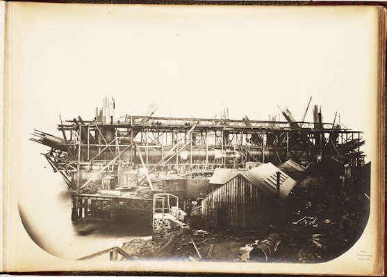 A large industrial structure under construction, featuring a complex framework of scaffolding and beams. Several cranes and wooden structures surround the area, and a body of water is visible beneath the unfinished framework.