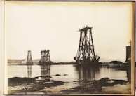 A vintage photograph depicts the early stages of massive bridge construction over a body of water. Several large, metal structures are partially built and tower over the surrounding landscape. The water in the foreground reflects the structures, while smaller cranes and equipment are visible. The background includes a shoreline with hills and a few buildings.