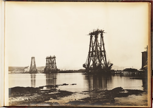A vintage photograph depicts the early stages of massive bridge construction over a body of water. Several large, metal structures are partially built and tower over the surrounding landscape. The water in the foreground reflects the structures, while smaller cranes and equipment are visible. The background includes a shoreline with hills and a few buildings.