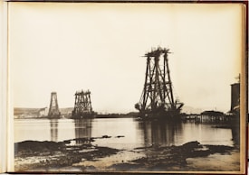 A vintage photograph depicts the early stages of massive bridge construction over a body of water. Several large, metal structures are partially built and tower over the surrounding landscape. The water in the foreground reflects the structures, while smaller cranes and equipment are visible. The background includes a shoreline with hills and a few buildings.