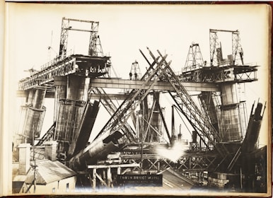 A black and white photograph showcasing the construction of a large steel bridge. The structure is partially built, with multiple steel beams and supports in various stages of assembly. Scaffolding and cranes are visible, indicating heavy industrial work. The bridge towers above a small building in the foreground, and there are several workers visible on the structure.