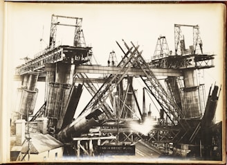 A black and white photograph showcasing the construction of a large steel bridge. The structure is partially built, with multiple steel beams and supports in various stages of assembly. Scaffolding and cranes are visible, indicating heavy industrial work. The bridge towers above a small building in the foreground, and there are several workers visible on the structure.