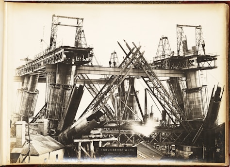 A black and white photograph showcasing the construction of a large steel bridge. The structure is partially built, with multiple steel beams and supports in various stages of assembly. Scaffolding and cranes are visible, indicating heavy industrial work. The bridge towers above a small building in the foreground, and there are several workers visible on the structure.