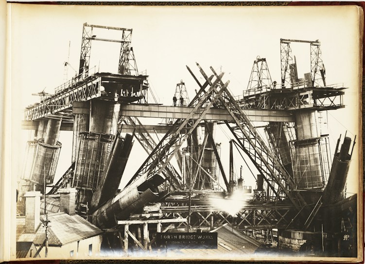 A black and white photograph showcasing the construction of a large steel bridge. The structure is partially built, with multiple steel beams and supports in various stages of assembly. Scaffolding and cranes are visible, indicating heavy industrial work. The bridge towers above a small building in the foreground, and there are several workers visible on the structure.