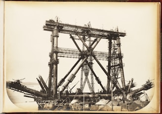 A large, complex metal structure, possibly part of a bridge under construction, dominates the scene. The framework consists of massive steel beams and girders arranged in a triangular pattern. The background includes a body of water and distant hills, while the foreground features various construction materials and small buildings.