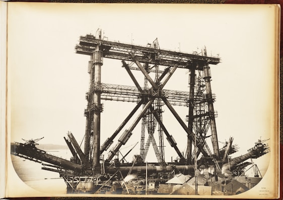 A large, complex metal structure, possibly part of a bridge under construction, dominates the scene. The framework consists of massive steel beams and girders arranged in a triangular pattern. The background includes a body of water and distant hills, while the foreground features various construction materials and small buildings.
