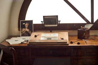A vintage wooden desk is situated by a window, featuring a portrait photograph, multiple drawing tools, and sheets of paper with sketches. An old-fashioned magnifying device is mounted on the desk, and a small wooden box holds brushes and pens.