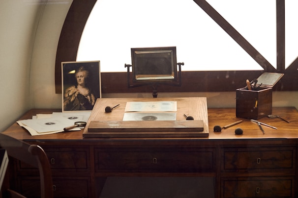 A vintage wooden desk is situated by a window, featuring a portrait photograph, multiple drawing tools, and sheets of paper with sketches. An old-fashioned magnifying device is mounted on the desk, and a small wooden box holds brushes and pens.