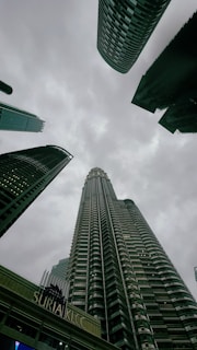 Tall skyscrapers reach up into a cloudy sky, with the Petronas Twin Towers prominently featured in the center. The architecture of the buildings is modern and sleek, and the skyline is composed of a mix of cylinder and rectangular shapes. The foreground shows an entrance to a shopping center labeled Suria KLCC, adding an urban commercial aspect to the scene.