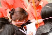 A group of people engaged in an activity, with several individuals wearing orange shirts. One person is holding a piece of paper and a pen, appearing to be focused on writing or reading. Colored ropes or strings are visible, crossing the scene diagonally.