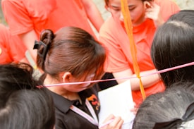 A group of people engaged in an activity, with several individuals wearing orange shirts. One person is holding a piece of paper and a pen, appearing to be focused on writing or reading. Colored ropes or strings are visible, crossing the scene diagonally.