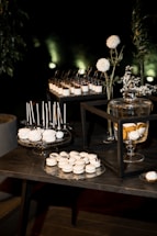 A dessert table arrangement features an array of sweets, including white macarons and cake pops displayed on decorative trays. Glass jars with a lemon-infused beverage are present, alongside vases with white flowers and greenery. The setting is elegant, with a dark background that highlights the illuminated desserts.