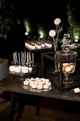 A dessert table arrangement features an array of sweets, including white macarons and cake pops displayed on decorative trays. Glass jars with a lemon-infused beverage are present, alongside vases with white flowers and greenery. The setting is elegant, with a dark background that highlights the illuminated desserts.