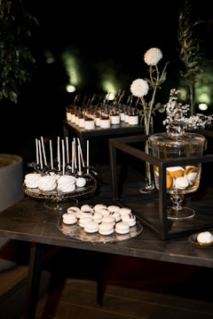 A dessert table arrangement features an array of sweets, including white macarons and cake pops displayed on decorative trays. Glass jars with a lemon-infused beverage are present, alongside vases with white flowers and greenery. The setting is elegant, with a dark background that highlights the illuminated desserts.