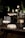 A dessert table arrangement features an array of sweets, including white macarons and cake pops displayed on decorative trays. Glass jars with a lemon-infused beverage are present, alongside vases with white flowers and greenery. The setting is elegant, with a dark background that highlights the illuminated desserts.
