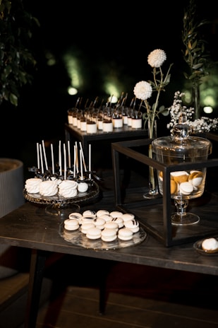 A dessert table arrangement features an array of sweets, including white macarons and cake pops displayed on decorative trays. Glass jars with a lemon-infused beverage are present, alongside vases with white flowers and greenery. The setting is elegant, with a dark background that highlights the illuminated desserts.