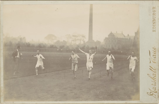 The image captures a historic footrace with five male runners competing. The participants, dressed in white athletic attire, are sprinting towards the finish line. One runner, with his arms raised in triumph, appears to be in the lead, suggesting a close finish. A man is standing nearby, possibly a race official or coach. The background features a grand, old building and a tall chimney or tower, partially obscured by trees, giving a sense of the event occurring in an older urban setting.