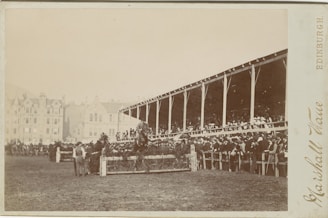 A large crowd gathers around a racetrack, with spectators seated in a wooden grandstand. In the foreground, a horse and rider are captured in mid-jump over a hurdle adorned with greenery. The background features a line of old buildings, adding a historic charm to the scene.