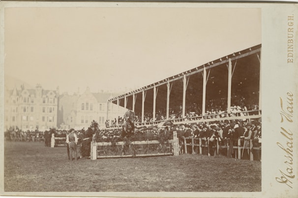 A large crowd gathers around a racetrack, with spectators seated in a wooden grandstand. In the foreground, a horse and rider are captured in mid-jump over a hurdle adorned with greenery. The background features a line of old buildings, adding a historic charm to the scene.