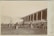 A large crowd gathers around a racetrack, with spectators seated in a wooden grandstand. In the foreground, a horse and rider are captured in mid-jump over a hurdle adorned with greenery. The background features a line of old buildings, adding a historic charm to the scene.