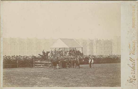 A historical photograph depicting a horse jumping over a fence in front of a crowd. The audience is gathered around a pavilion, watching the event. The background features a row of old buildings, possibly in a city setting.