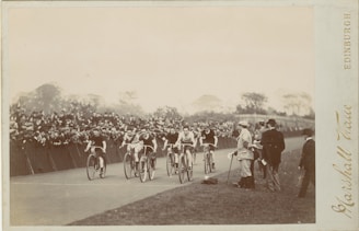 A group of cyclists are racing on a track with a crowd of spectators watching from the side. The cyclists are in action, with some standing on the side, possibly officials or team members. The scene appears vintage with muted colors and the photograph is likely from an older time period given the attire of the people and the style of the bicycles.