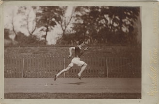 A monochrome photograph capturing a man in mid-sprint on an outdoor track. He is wearing a dark sleeveless top and light-colored shorts. The background shows a wooden fence and blurred, leafy trees.